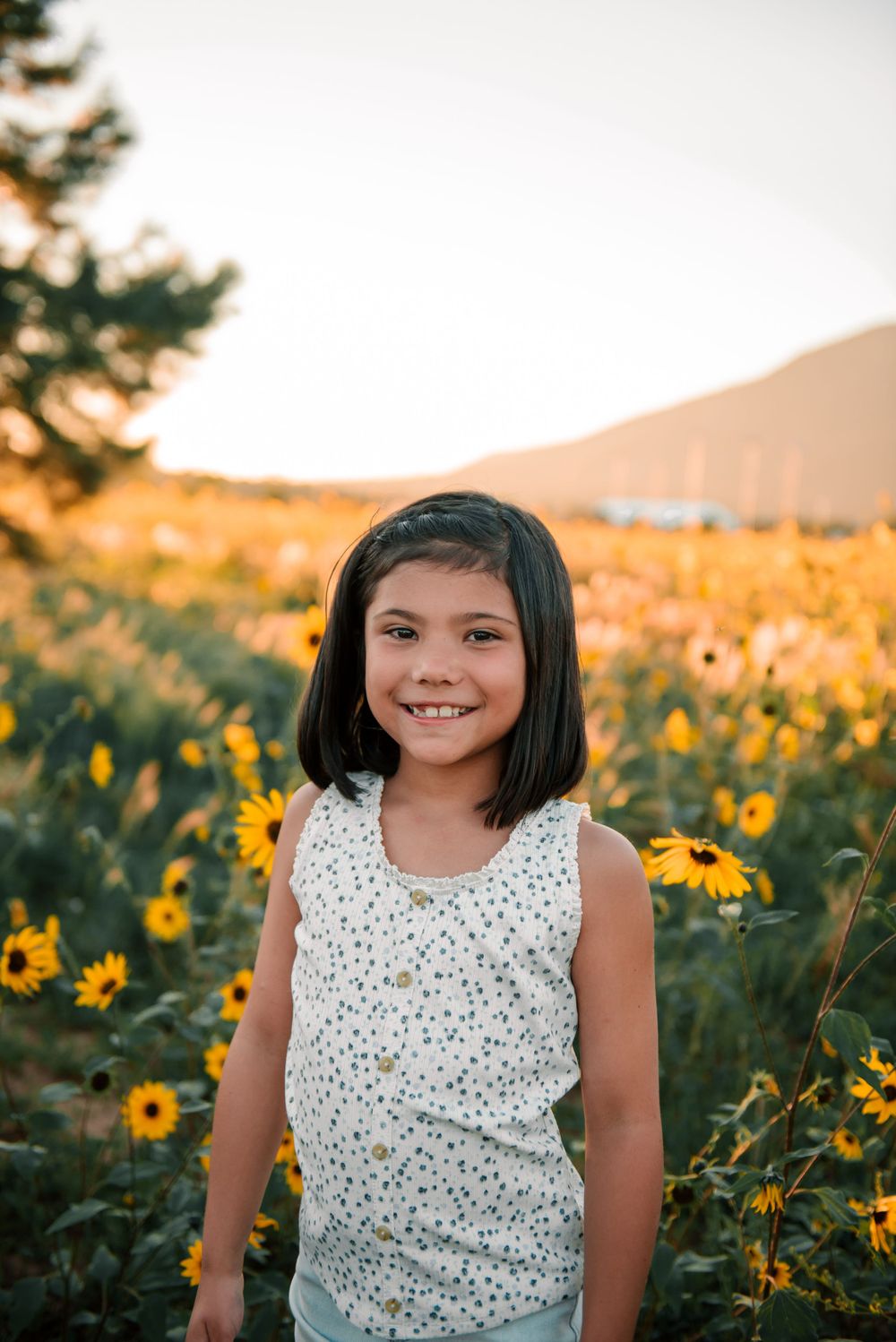 A child poses cheerfully in a floral top surrounded by bright sunflowers at sunset.