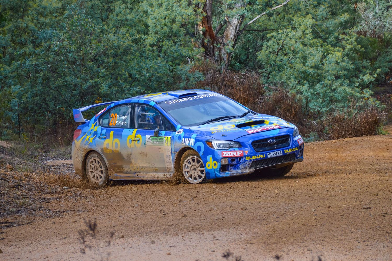 Blue Subaru rally car drifting through muddy corner on dirt course.