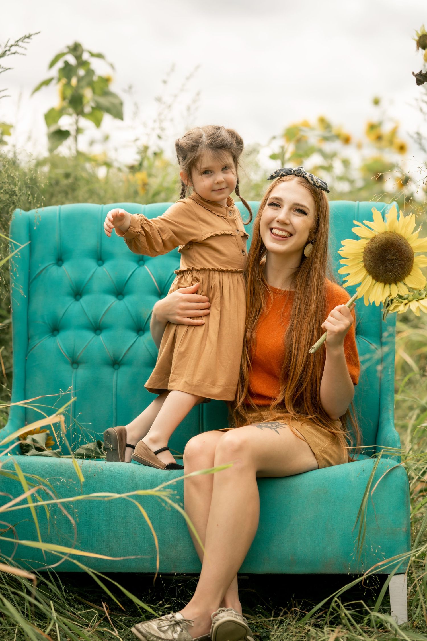 Mother and child sitting on a teal couch in a sunflower field during golden hour.
