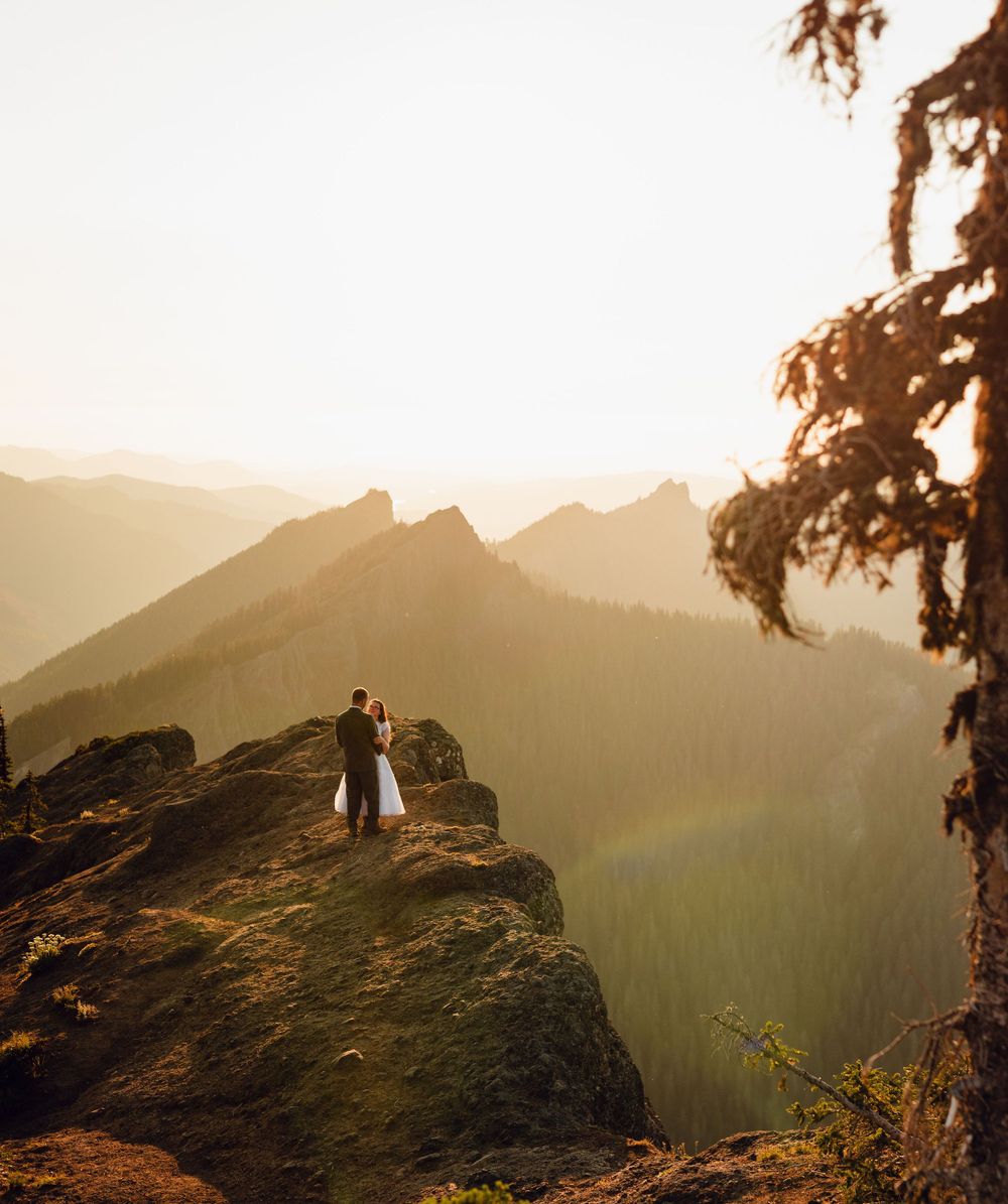 Newlyweds embrace on a mossy mountain peak surrounded by golden glowing forested mountain ranges.