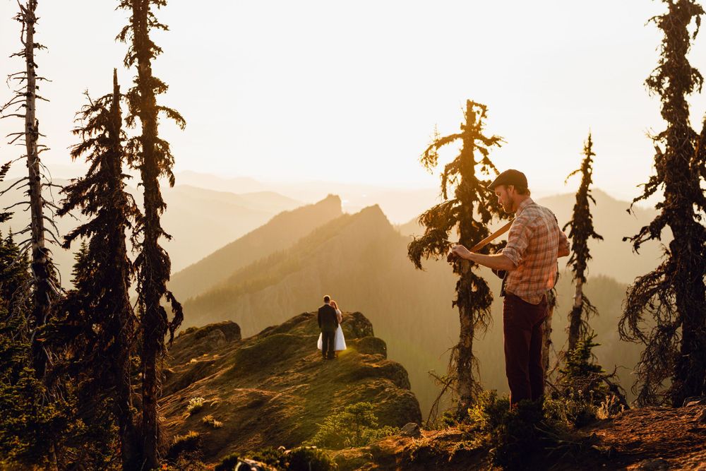 Hiker examines a small tree on a rocky mountain summit at golden hour with scenic peaks in the background.