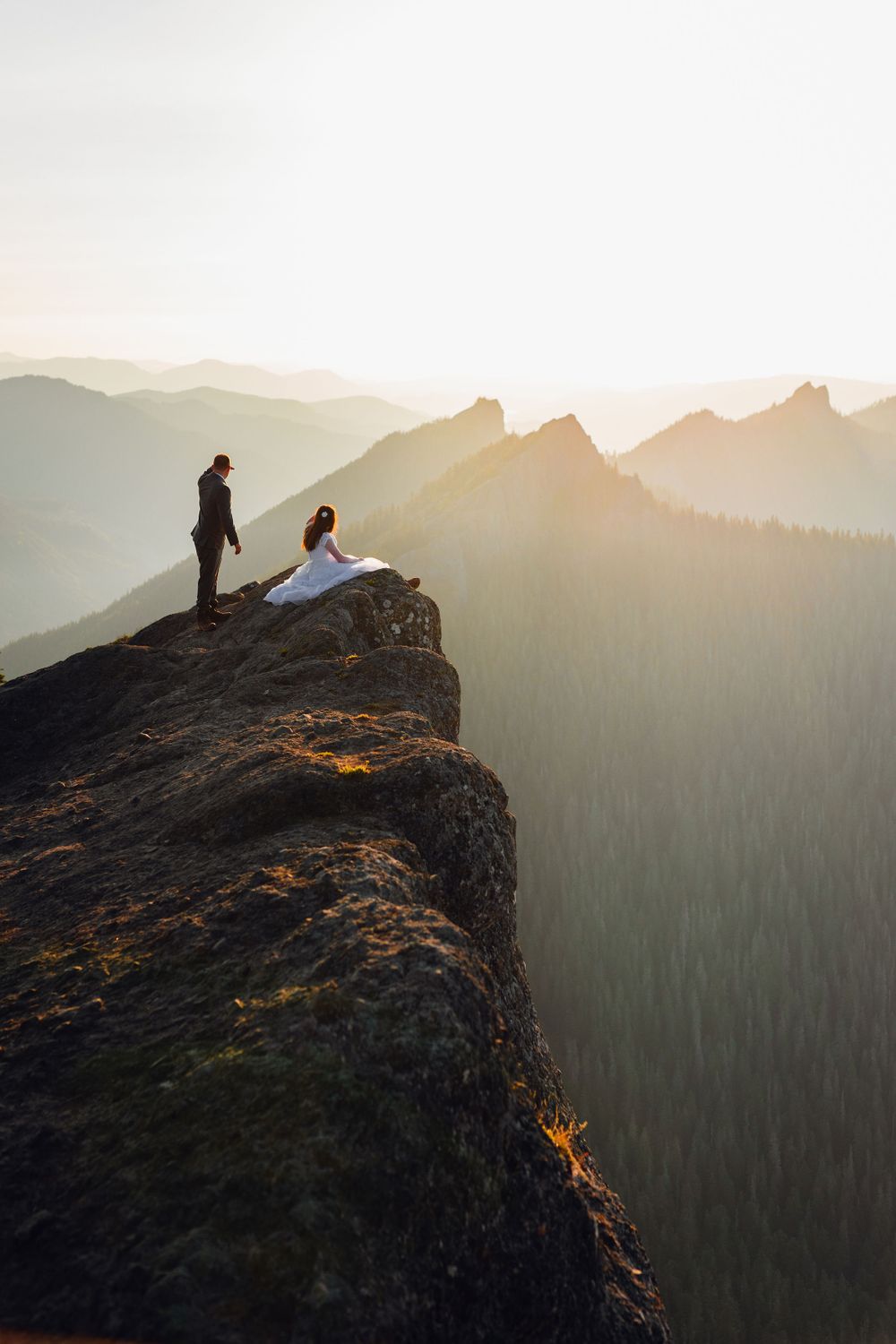 Bride and groom hold hands on a dramatic rocky cliff edge with layered mountain sunset backdrop.