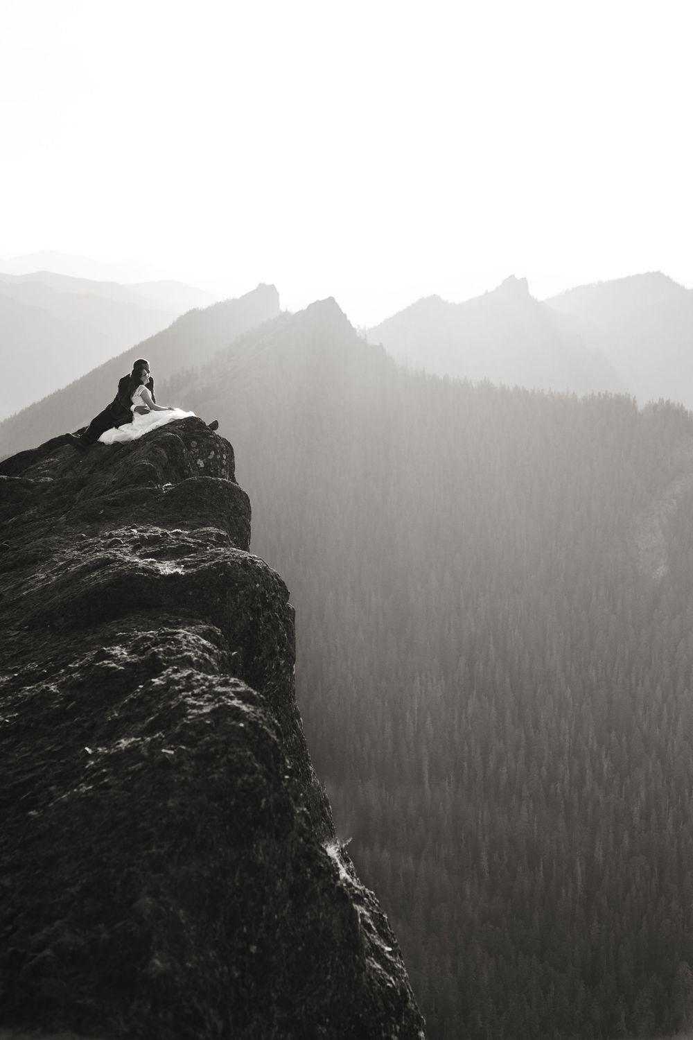Black and white photo of a bride sitting on a dramatic rocky cliff edge overlooking misty forested mountains.