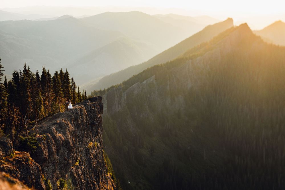 Tiny wedding couple stands on a rocky mountain cliff bathed in warm golden sunset light with layered peaks.
