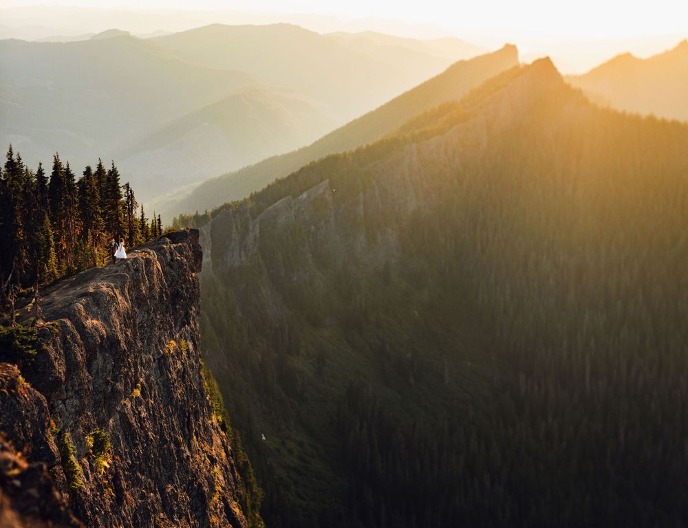 Distant wedding couple stands on a rugged mountain cliff edge surrounded by golden sunset and layered peaks.