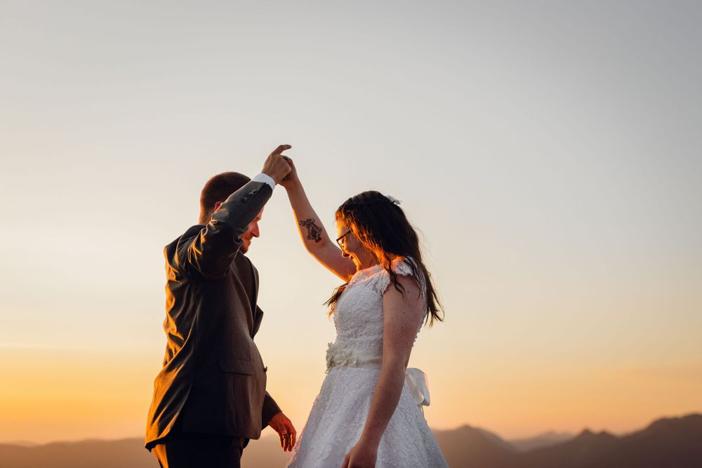 Bride and groom dance joyfully against a vibrant orange sunset sky with mountain silhouettes below.