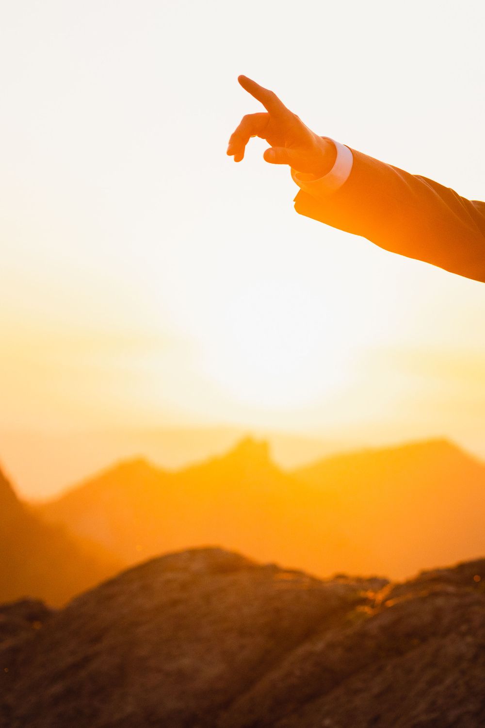 Close-up of a hand pointing against a vivid orange mountain sunset sky with a wedding ring visible.