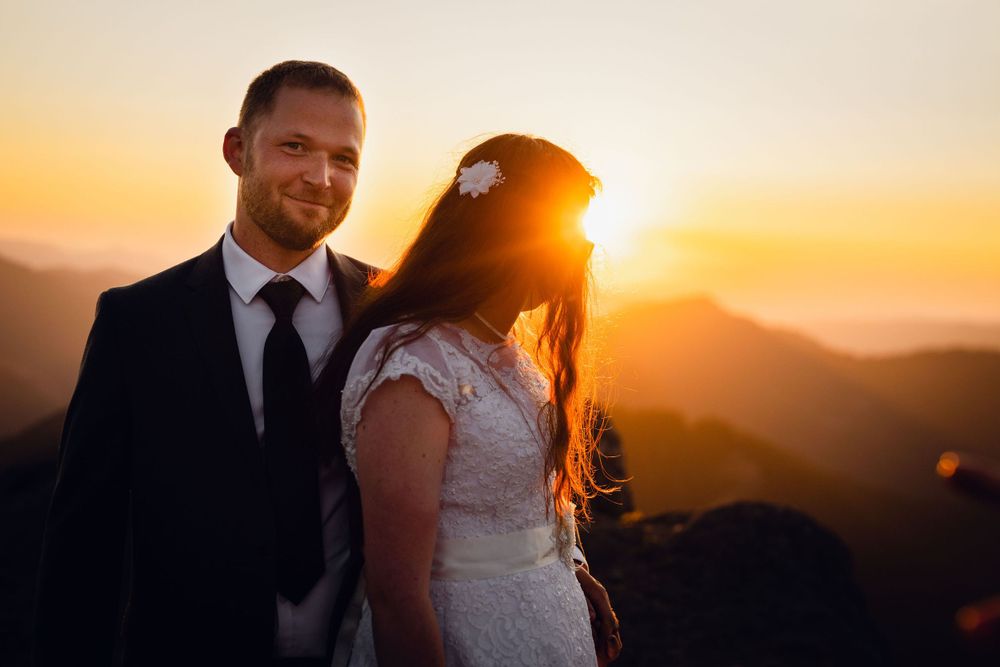 Smiling groom and bride pose together on a mountain top with a stunning golden sunset glowing behind her.