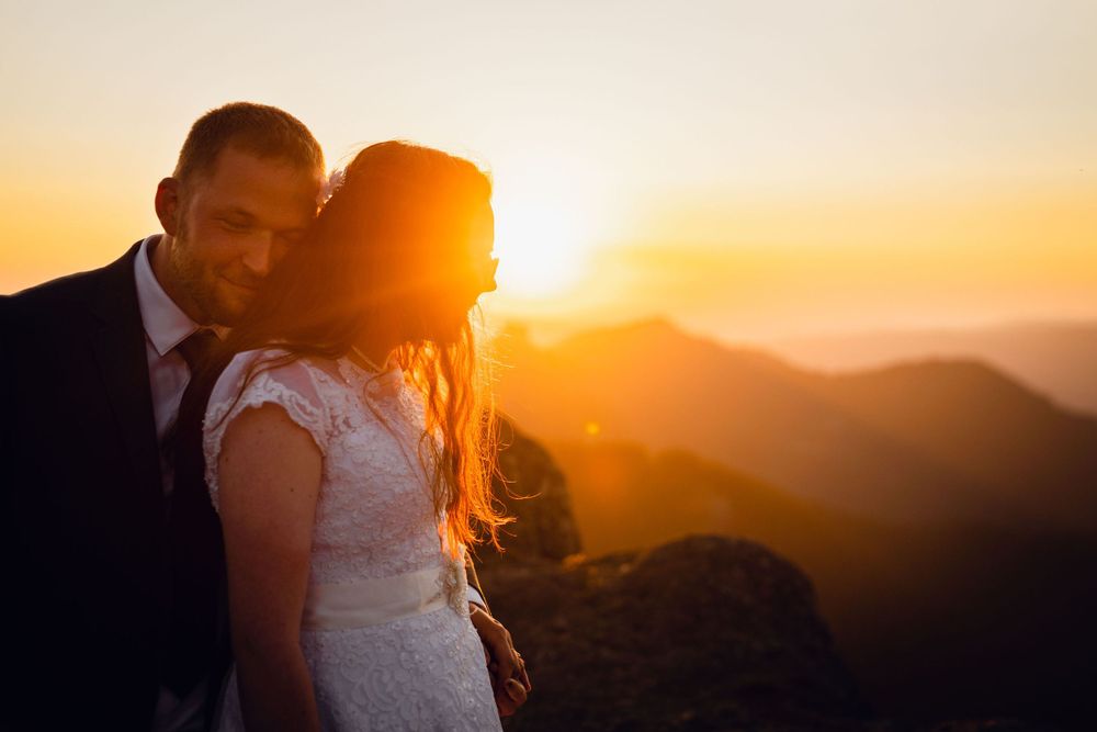 Bride and groom embrace tenderly on a rocky mountain summit with a glowing golden sunset behind them.