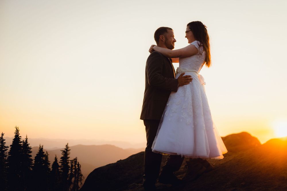 Bride in white dress and groom embracing on a mountain peak at golden sunset.