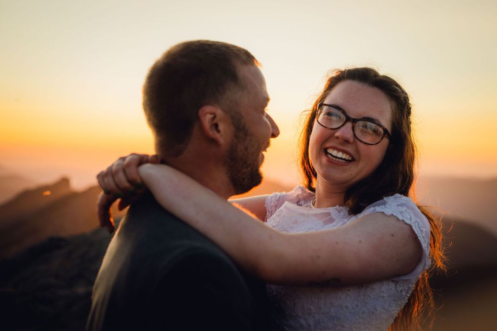 Smiling bride with glasses laughing joyfully with groom during sunset mountain elopement.
