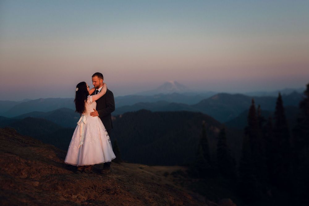 Wedding couple embracing on a mountain ridge at dusk with layered blue hills behind.