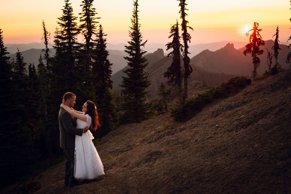 Couple in wedding attire embracing on a hillside surrounded by pine trees at sunset.
