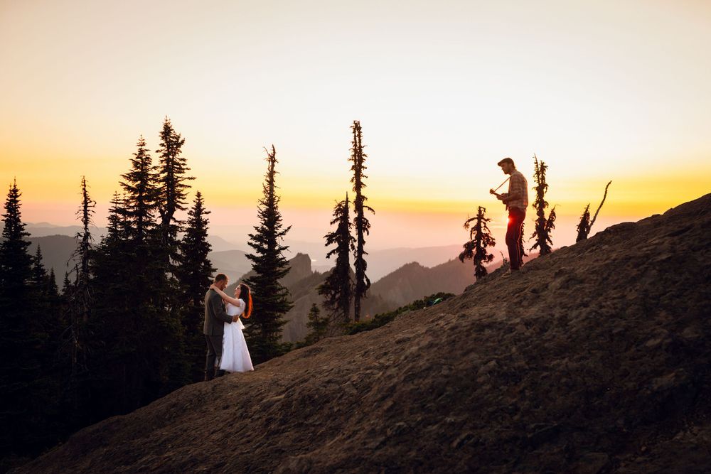 Couple elopes at sunset on a mountain hillside with evergreen trees silhouetted against a golden sky.