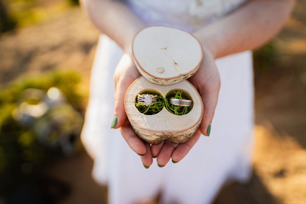 Bride holds a rustic wooden ring box containing two wedding bands with green moss detail.