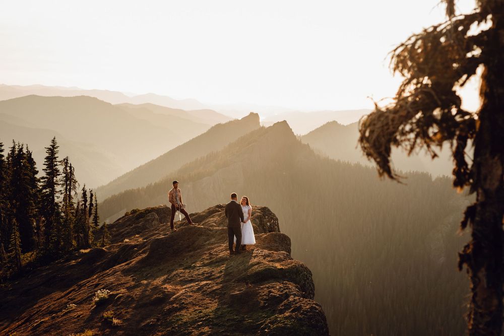 Wedding couple stands on a rocky mountain summit surrounded by golden sunset light and forested valleys.