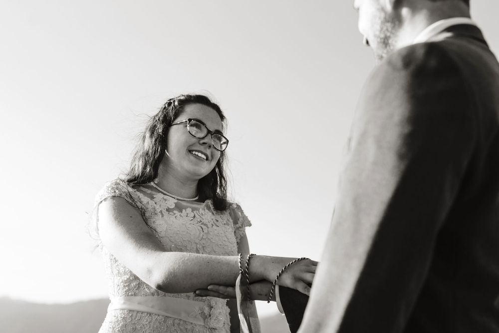 Black and white photo of a smiling bride with glasses holding hands during an outdoor wedding ceremony.