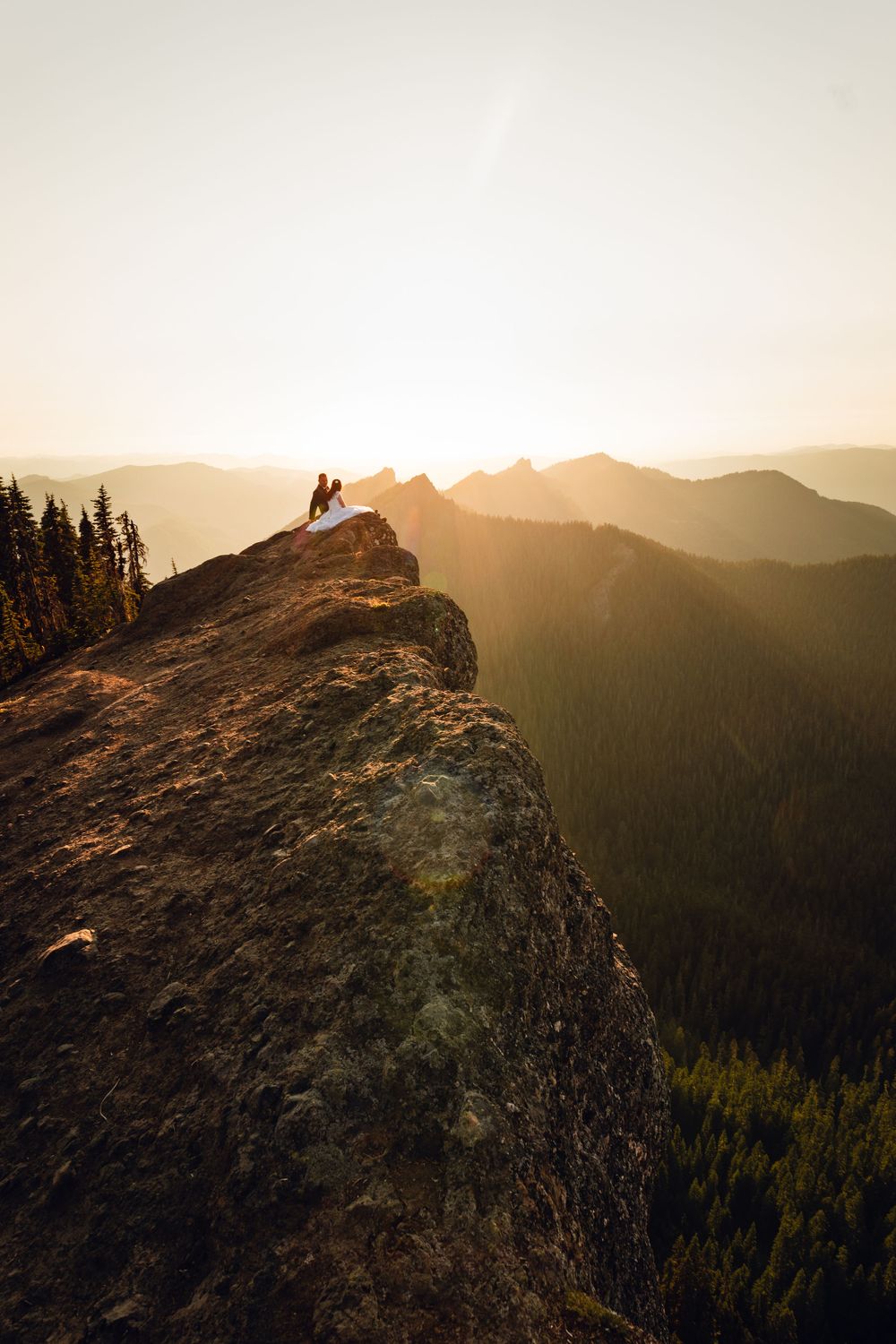 Couple sits together on a dramatic rocky mountain peak bathed in warm golden sunset light.