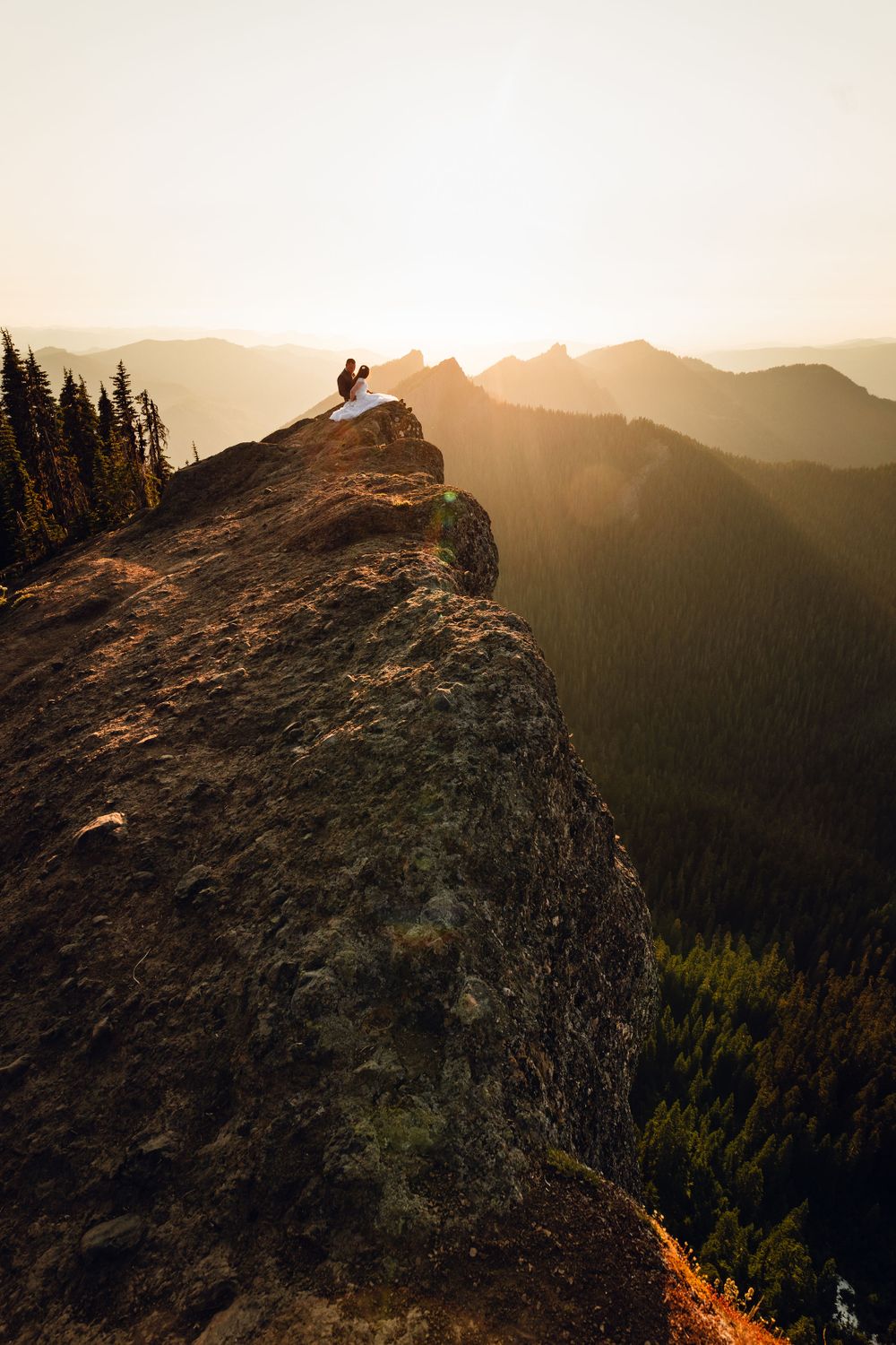 Wedding couple perched on a rugged mountain cliff edge with breathtaking sunset valley views below.