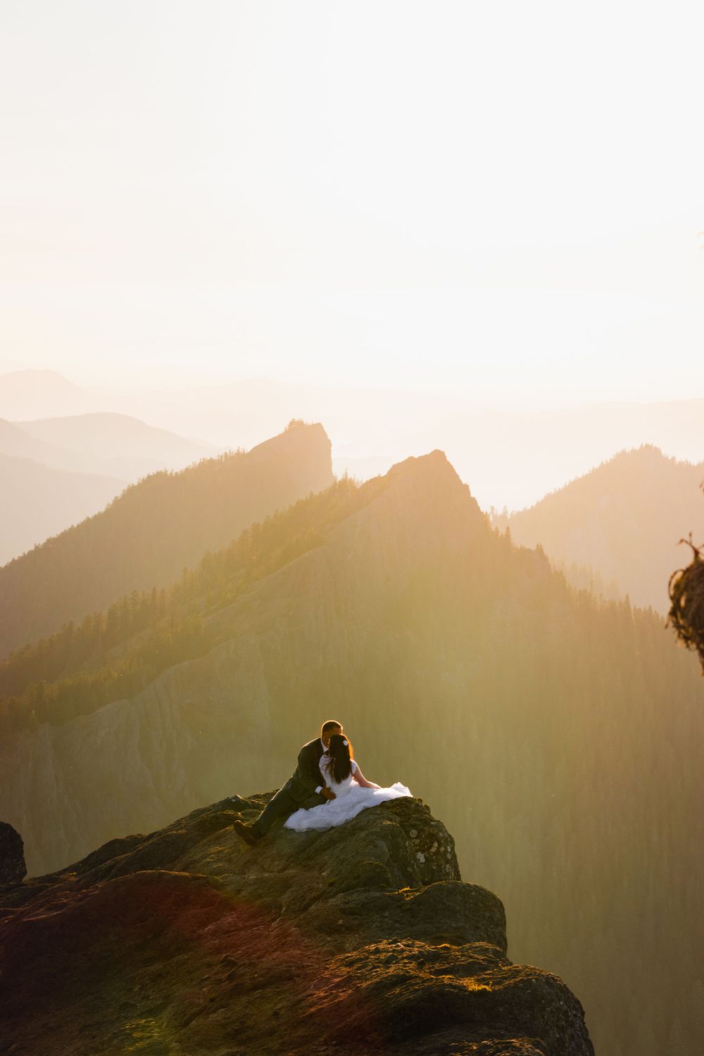 Bride and groom embrace on a rocky mountain summit with layered golden mountain ranges behind them.