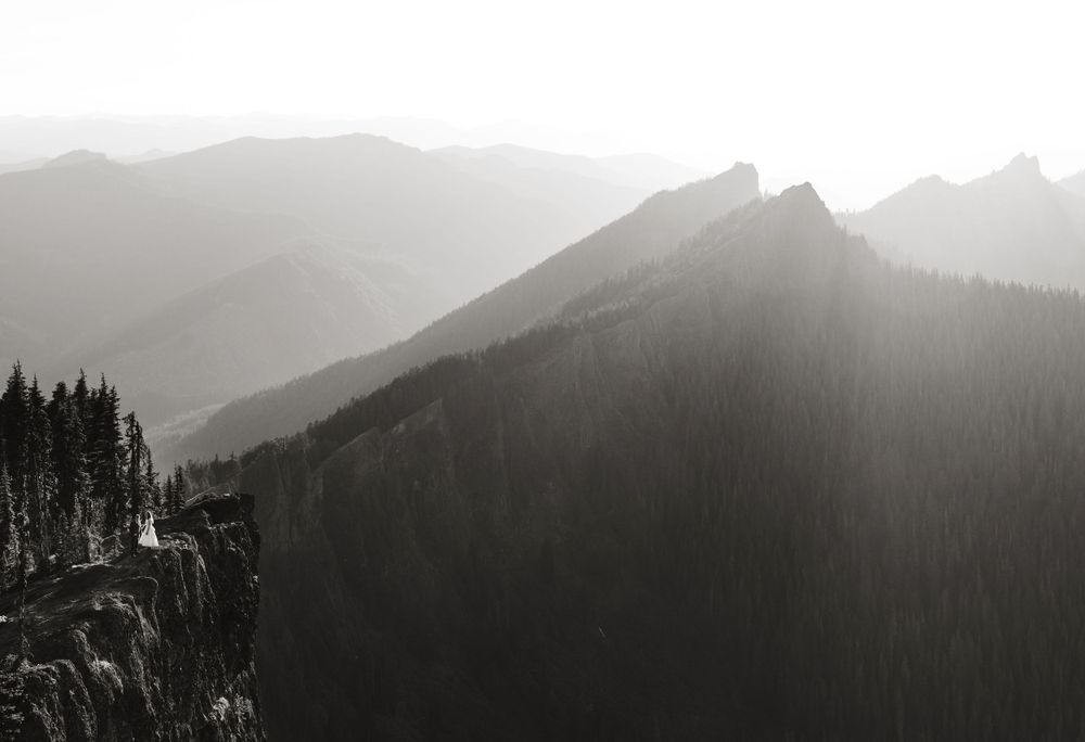Black and white aerial view of tiny wedding couple on a cliff edge amid misty mountain ranges.