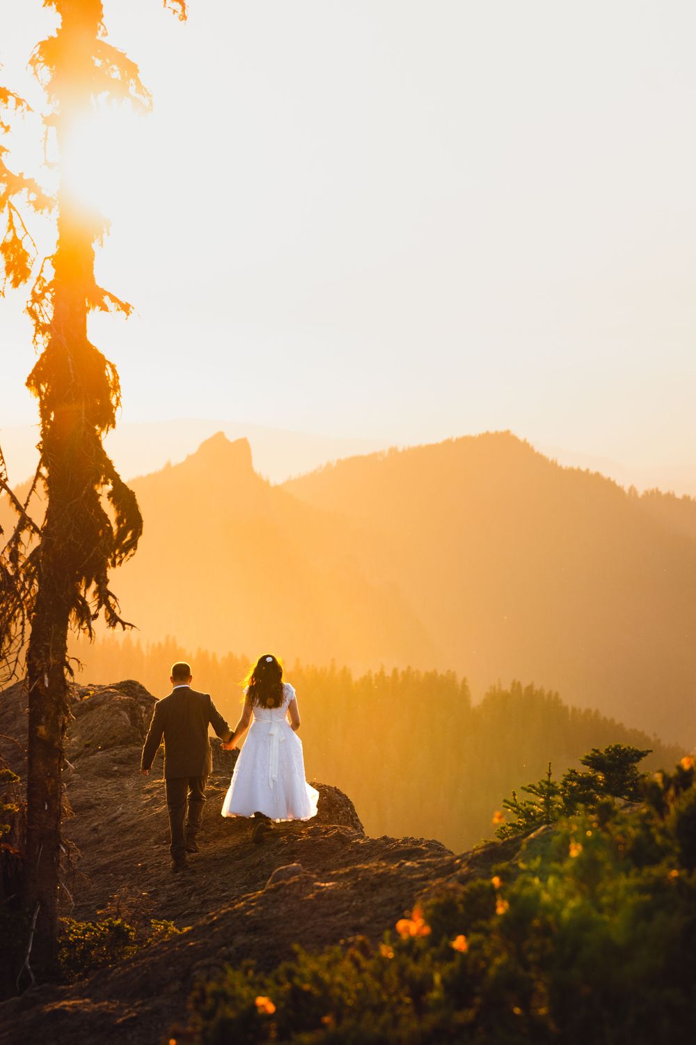 Bride in white gown and groom walking hand in hand on mountain ridge at sunset.