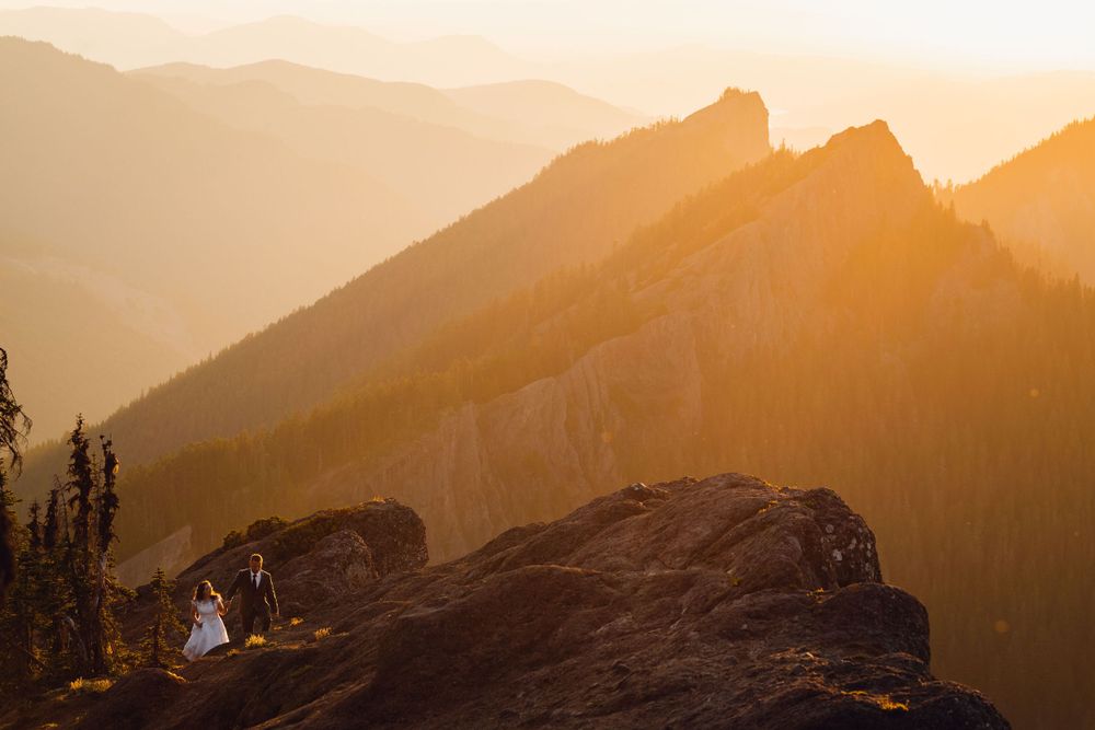 Wedding couple on a dramatic rocky cliff at golden sunset with layered mountain views.