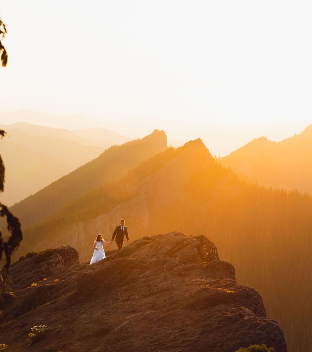 Couple in wedding attire standing on a rocky mountain summit during a glowing sunset.