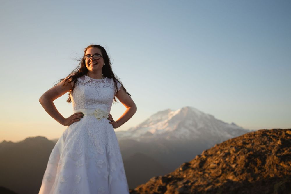 Smiling bride in lace wedding gown posing confidently with Mount Rainier at sunset.