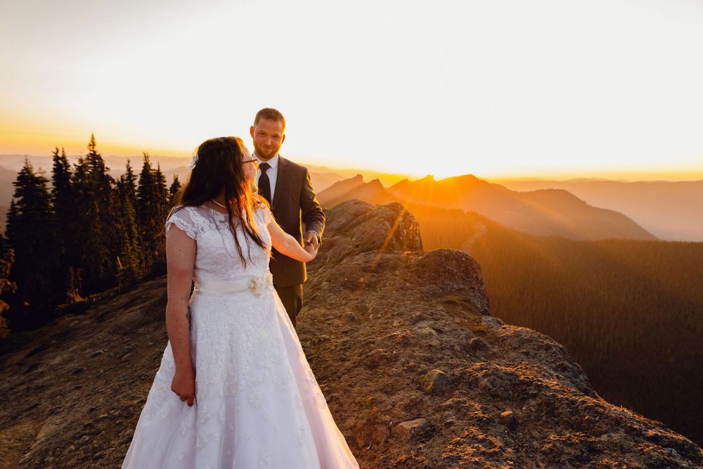 Bride and groom holding hands on a mountain ridge with a vibrant golden sunset behind.