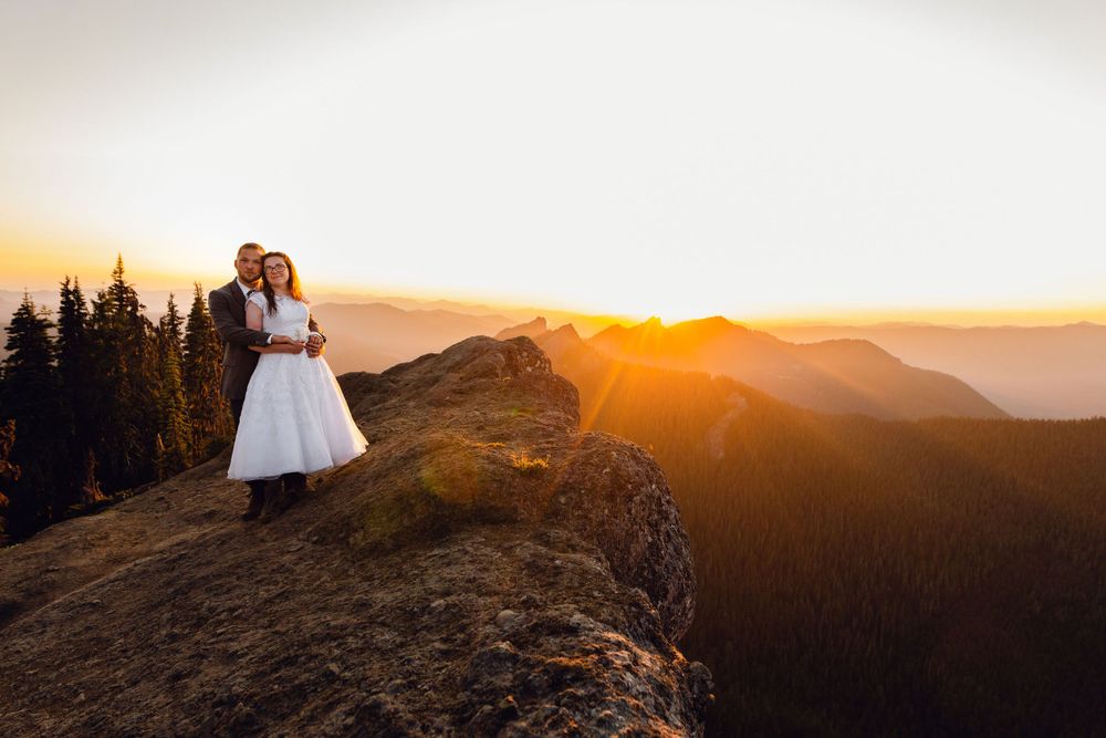 Bride and groom embrace on mountain summit at golden sunset with forest and mountain range backdrop.