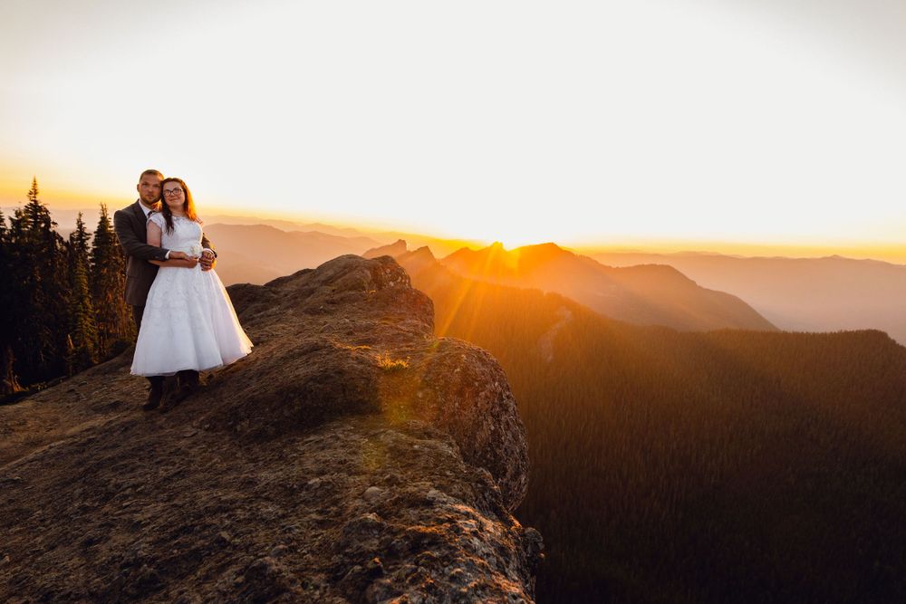 Wedding couple stands on rocky mountain peak during stunning golden hour sunset with layered mountains.