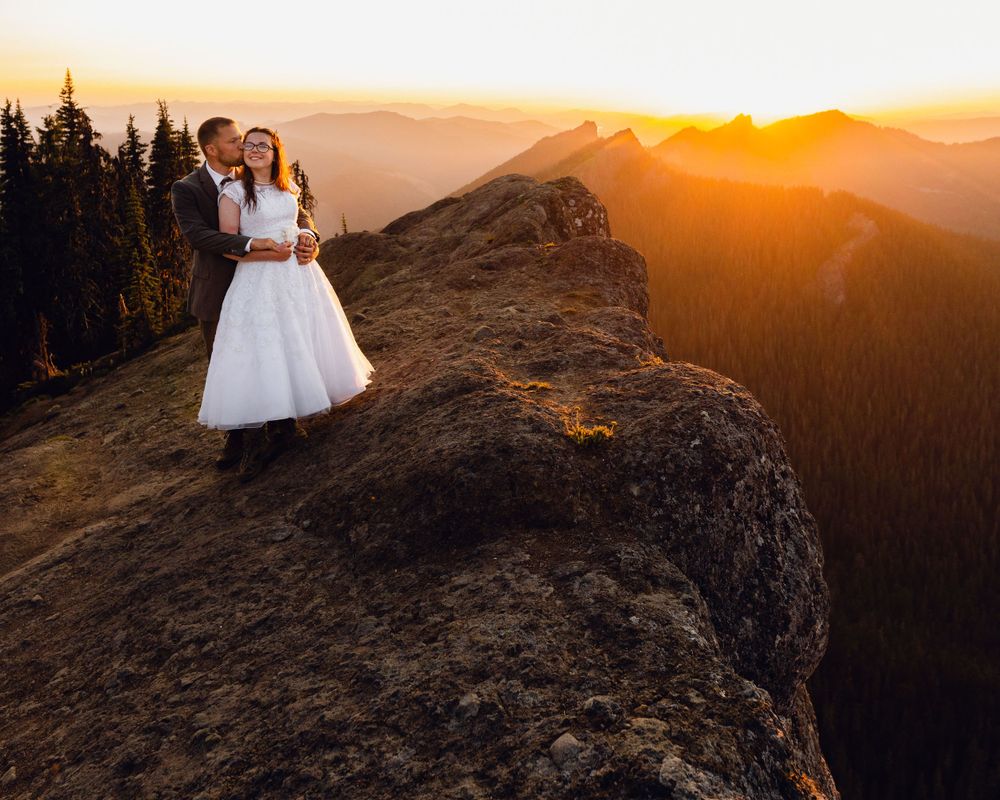 Groom kisses bride on rocky mountain summit with breathtaking golden sunset and evergreen forest view.