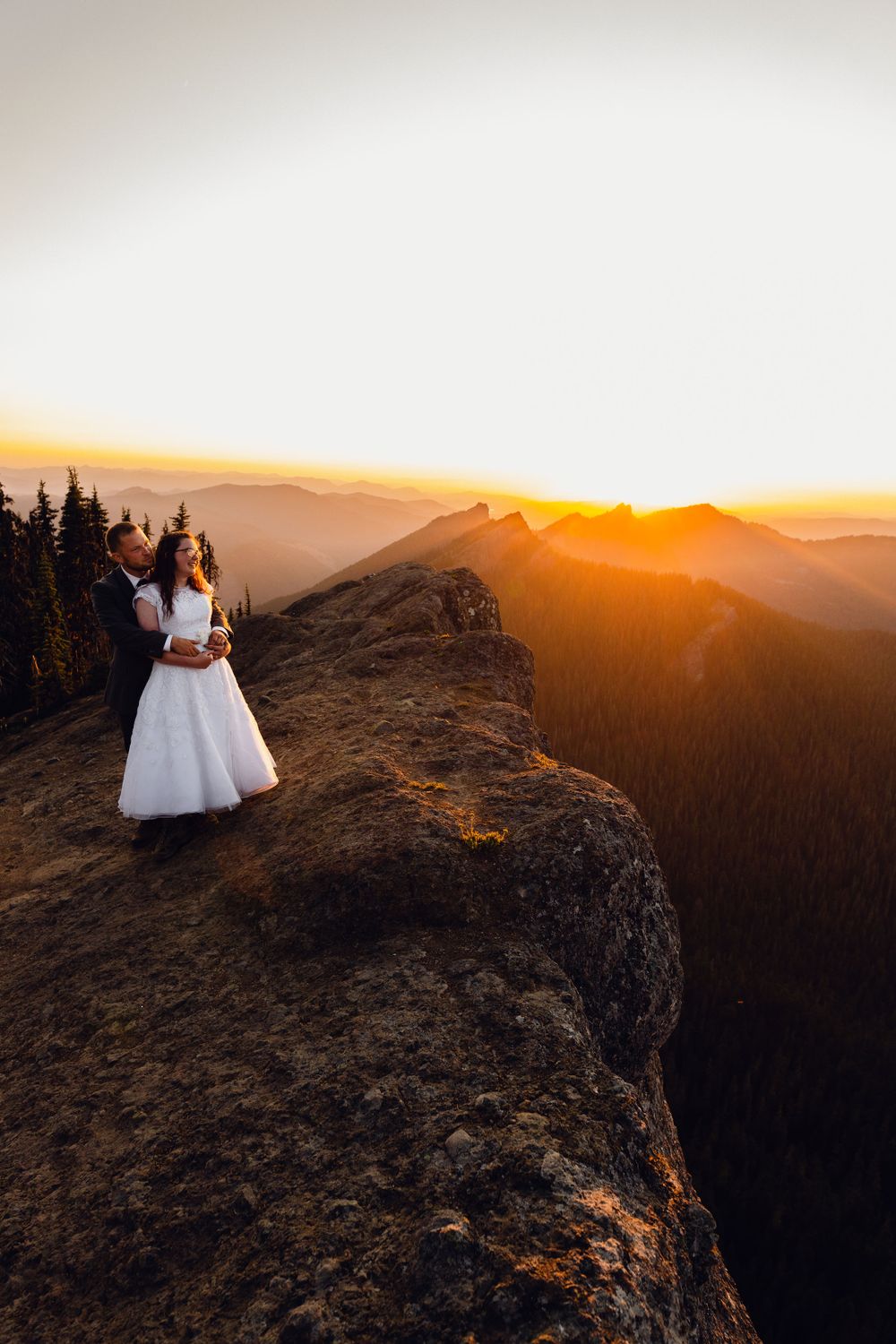Bride in white dress and groom stand on dramatic mountain cliff edge during vibrant orange sunset.