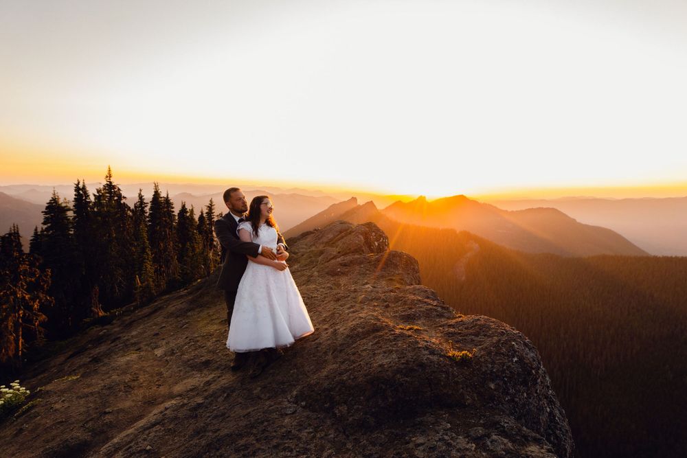 Romantic couple in wedding attire embraces on mountain peak with glowing sunset and misty valley below.