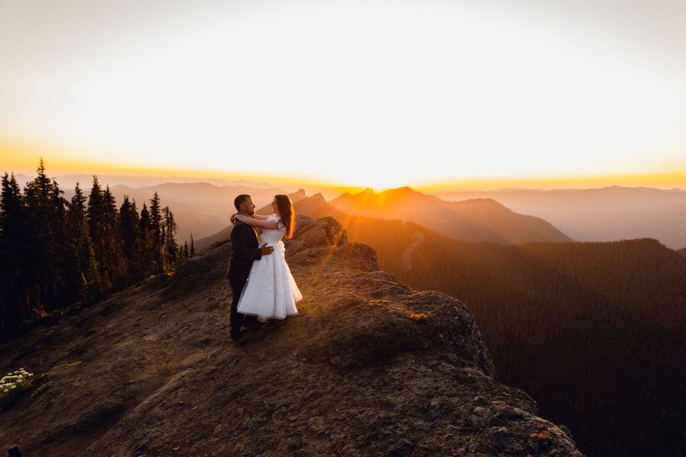 Bride and groom share joyful moment on mountain summit with brilliant golden sunset behind them.