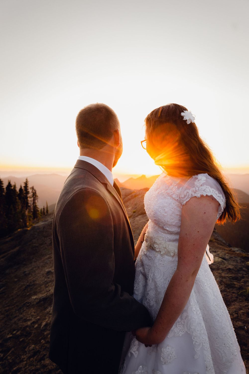 Wedding couple holds hands facing each other with sun flare between them at mountain sunset.