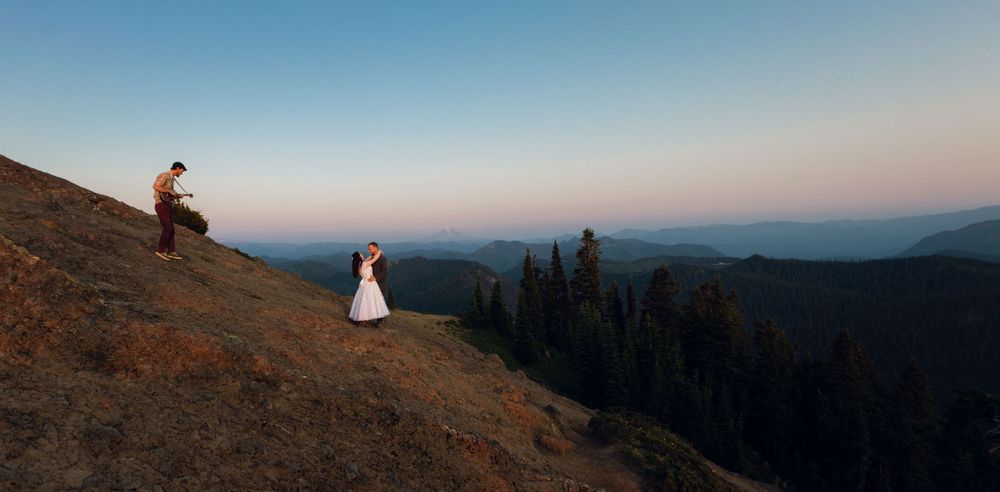 Wedding couple stands apart on steep mountain slope at dusk with vast mountain landscape behind.