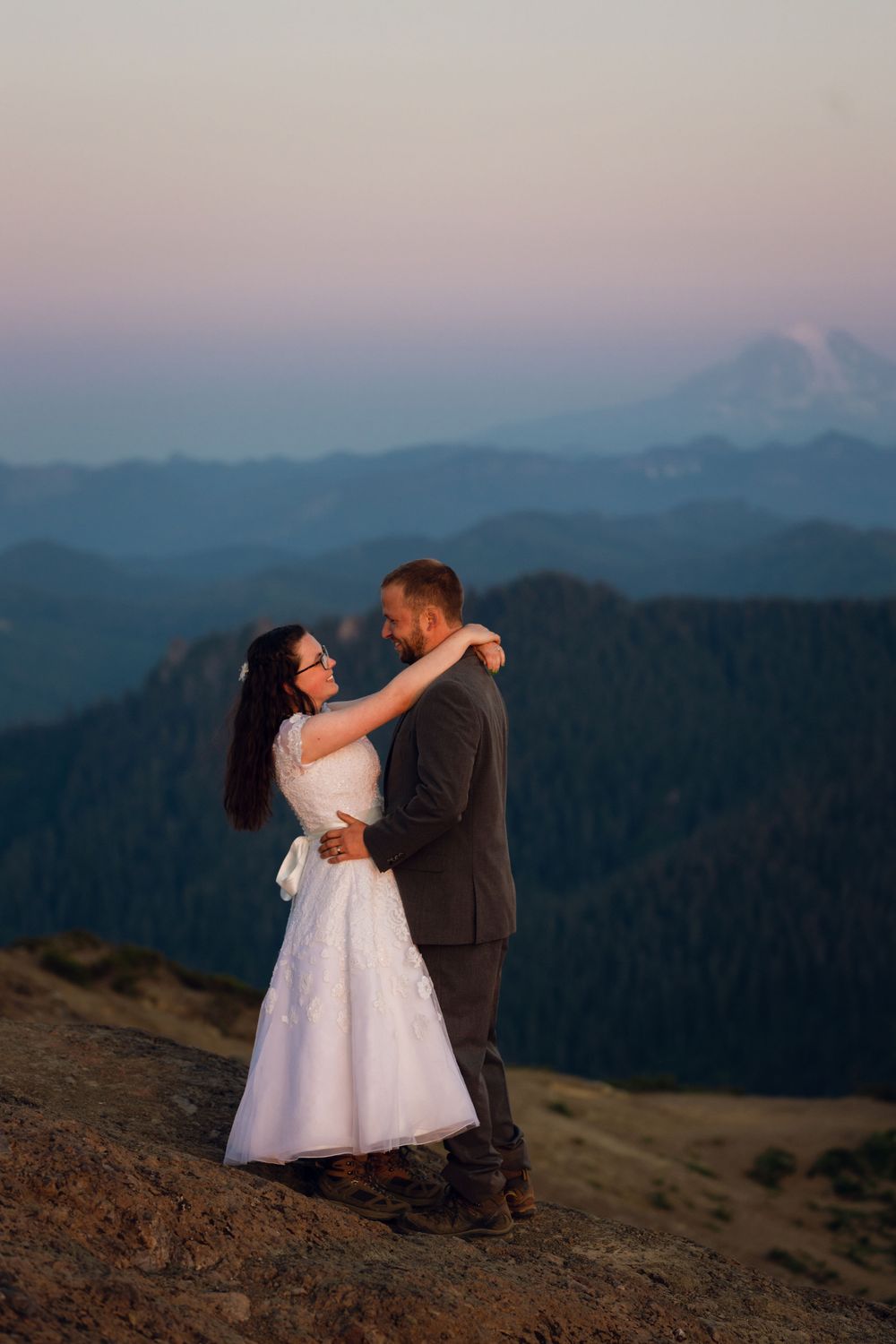 Bride and groom embrace on mountain summit at twilight with purple sky and distant snow-capped peak.