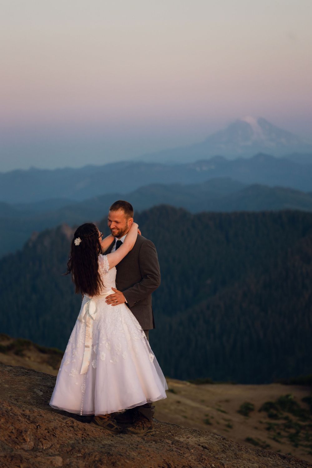 Bride and groom embrace on mountain summit at dusk with snow-capped peak and layered blue ridges behind them.
