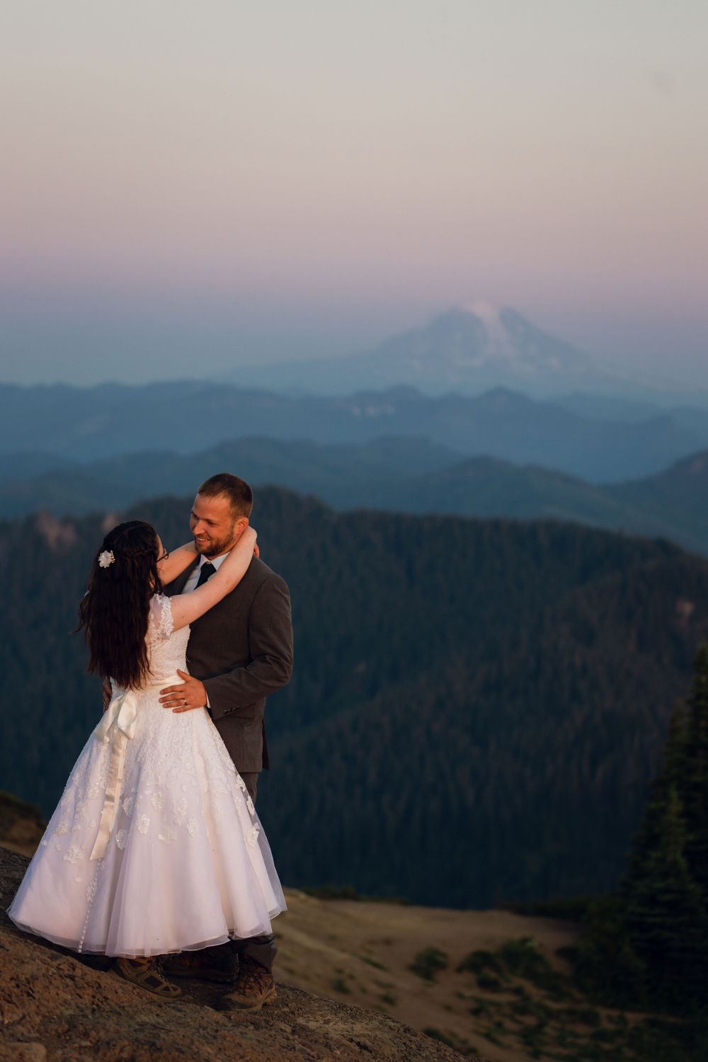 Wedding couple share romantic moment on rocky mountain overlook with misty blue peaks and snow-capped summit at sunset.