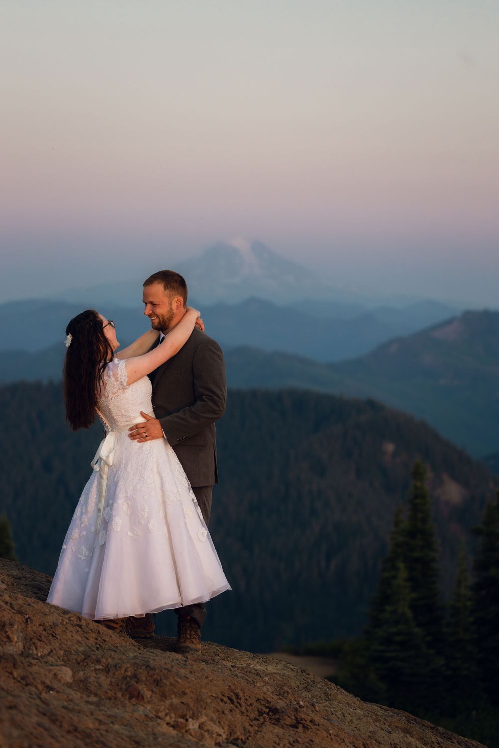 Smiling newlyweds embrace on mountain peak at twilight with snow-capped volcano and blue layered ridgelines in background.