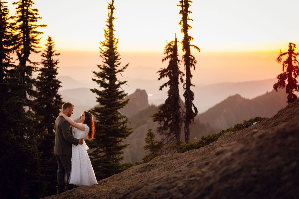 Bride and groom kiss on alpine hillside surrounded by tall evergreen trees with golden sunset over mountain valleys.