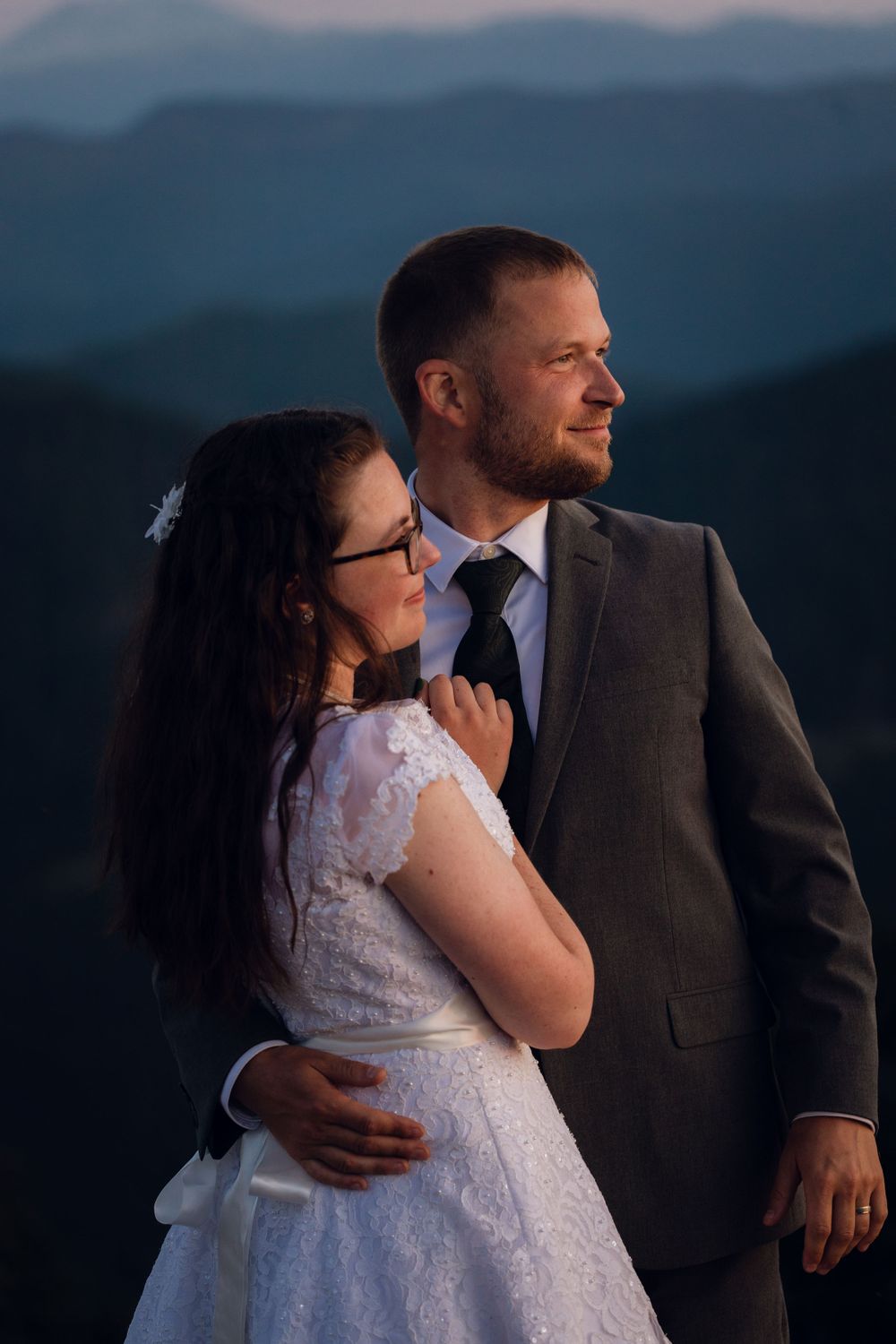 Newlywed couple stand together gazing into distance with dramatic blue mountain layers behind them at dusk.