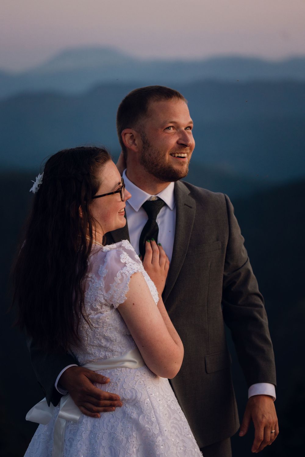 Bride in lace dress holds grooms tie as he smiles against a dusky blue mountain backdrop at sunset.