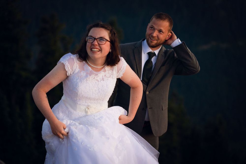 Laughing bride in full white gown and groom share a playful moment outdoors with dark evergreen trees behind.