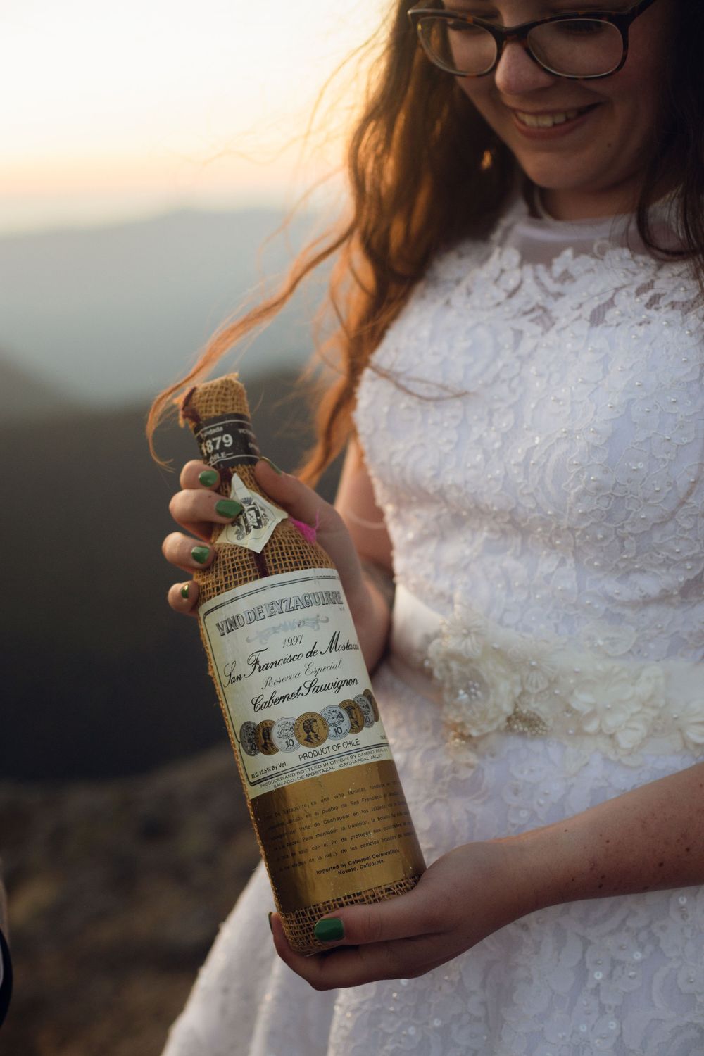 Smiling bride in lace wedding dress holds an antique wine bottle with mountain sunset glowing in the background.