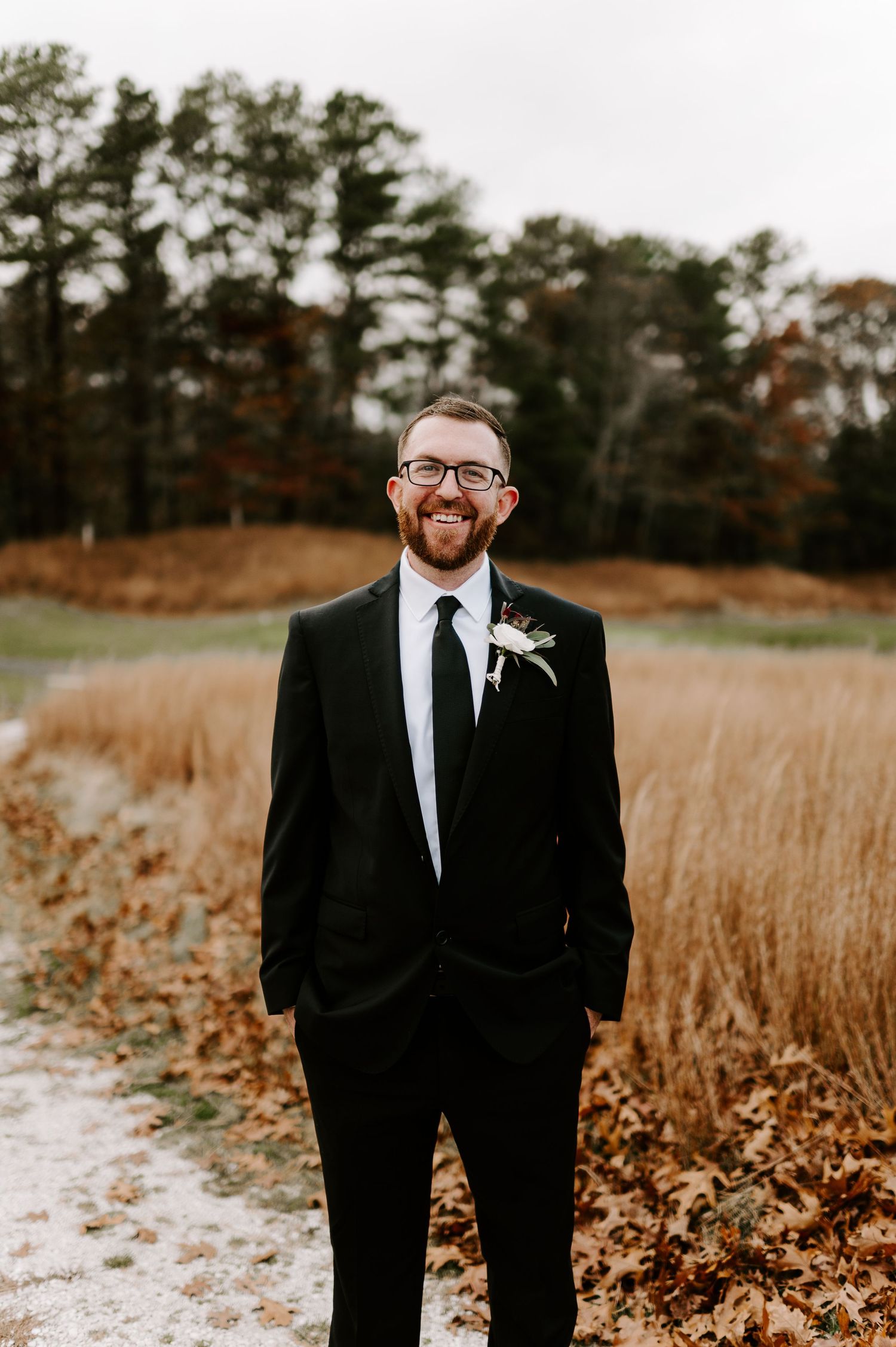 Groomsman in black suit and glasses poses in autumn field with golden grass.