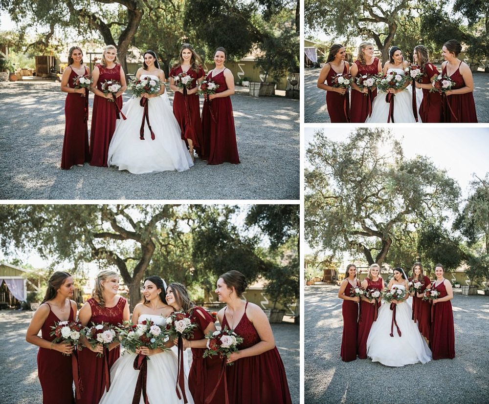Wedding party in burgundy bridesmaid dresses and white bridal gown pose together under oak trees at outdoor venue.