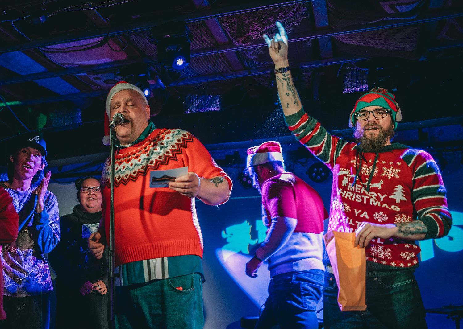 Musicians performing on stage wearing festive holiday sweaters during a concert with blue lighting effects.